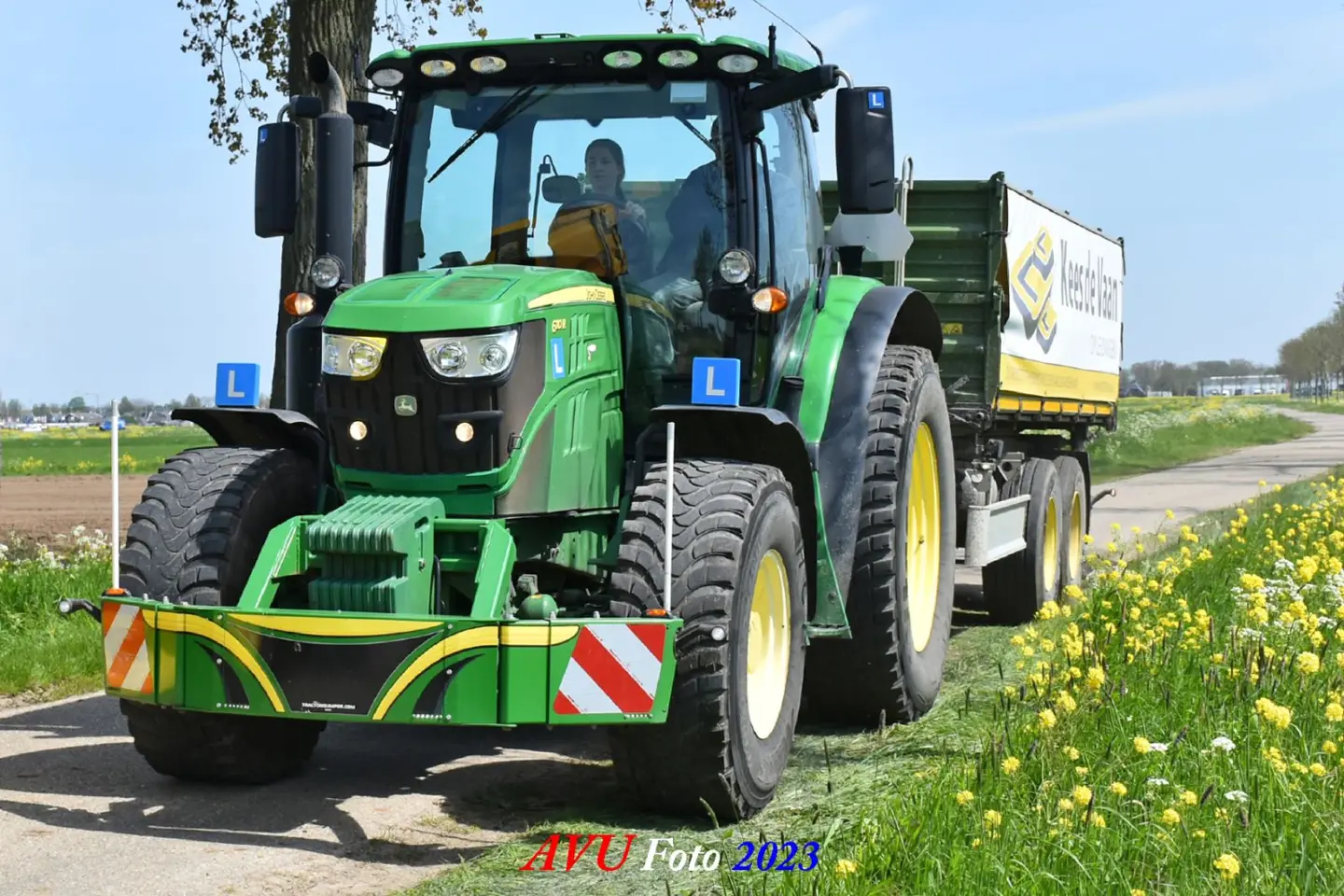 A green tractor fitted with a Box 50L without front linkage bumper is pulling a green trailer on a rural road