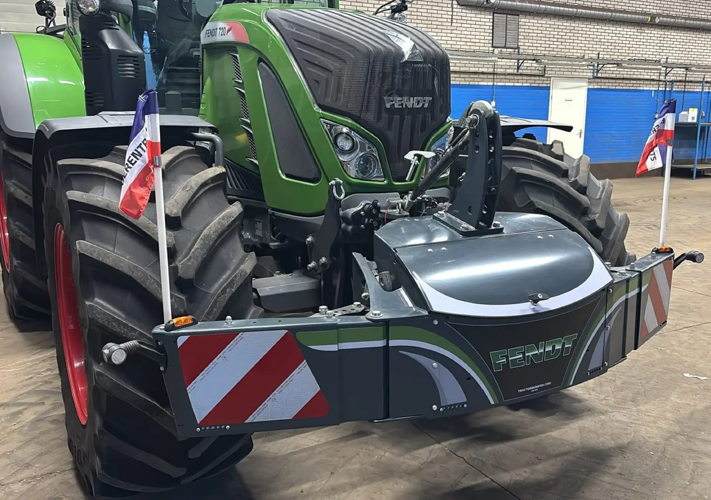 A tractor with a front weight bumper displaying Boerentrots protest flags on each side indoors.