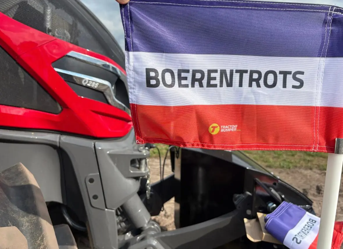 A hand holds a Boerentrots protest flag in front of a red and grey tractor with visible front weight bumper parts