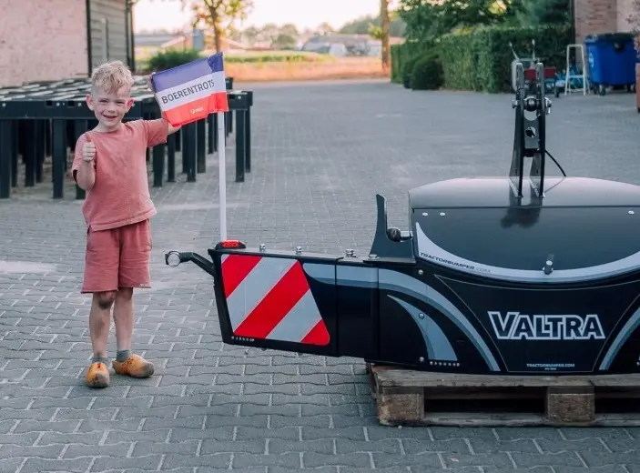 Smiling child holding a Boerentrots protest flag next to a large black front bumper with red and white stripes on a pallet