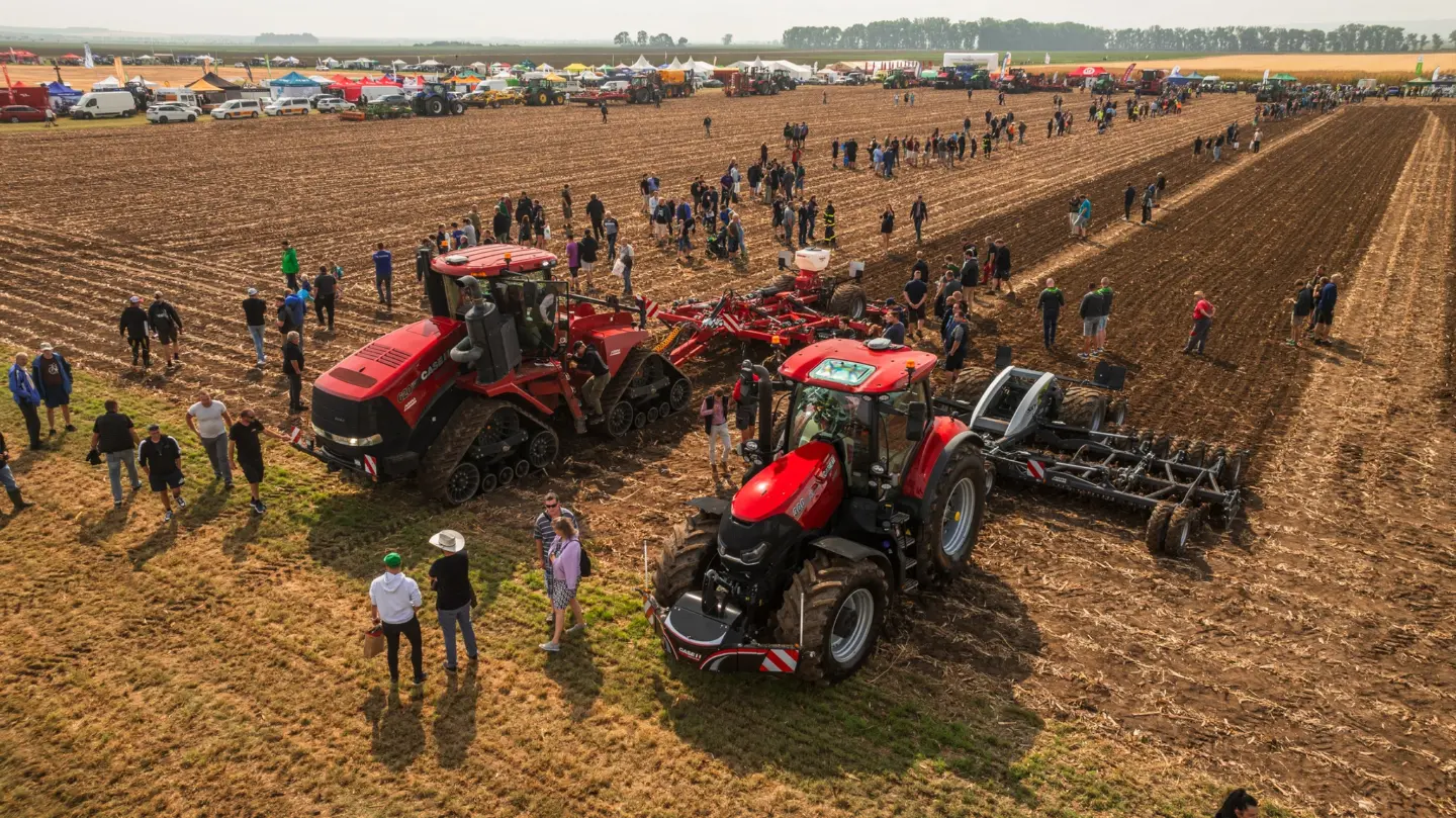 Two red tractors with front weight bumpers and implements are displayed in a field among groups of people and tents
