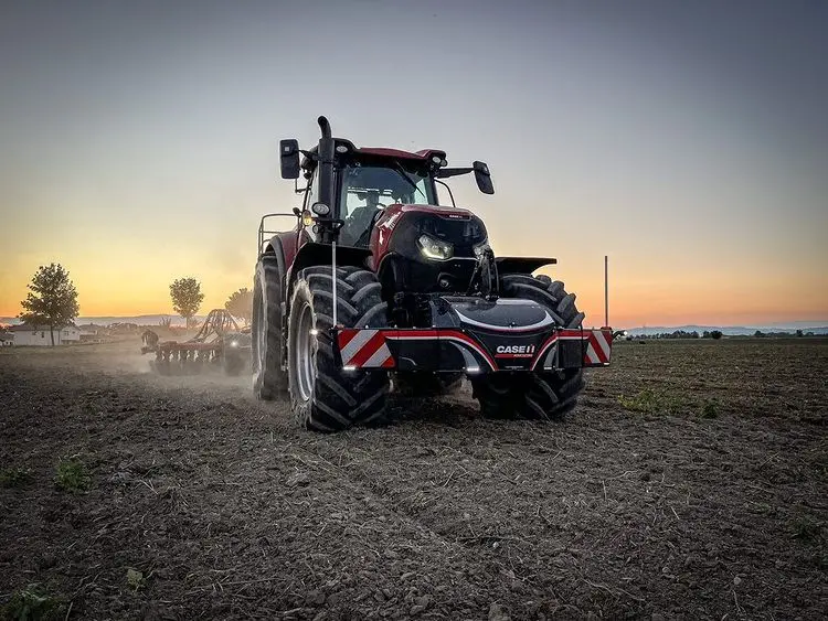 Red tractor with a front weight bumper featuring red and white stripes working in a field at sunset