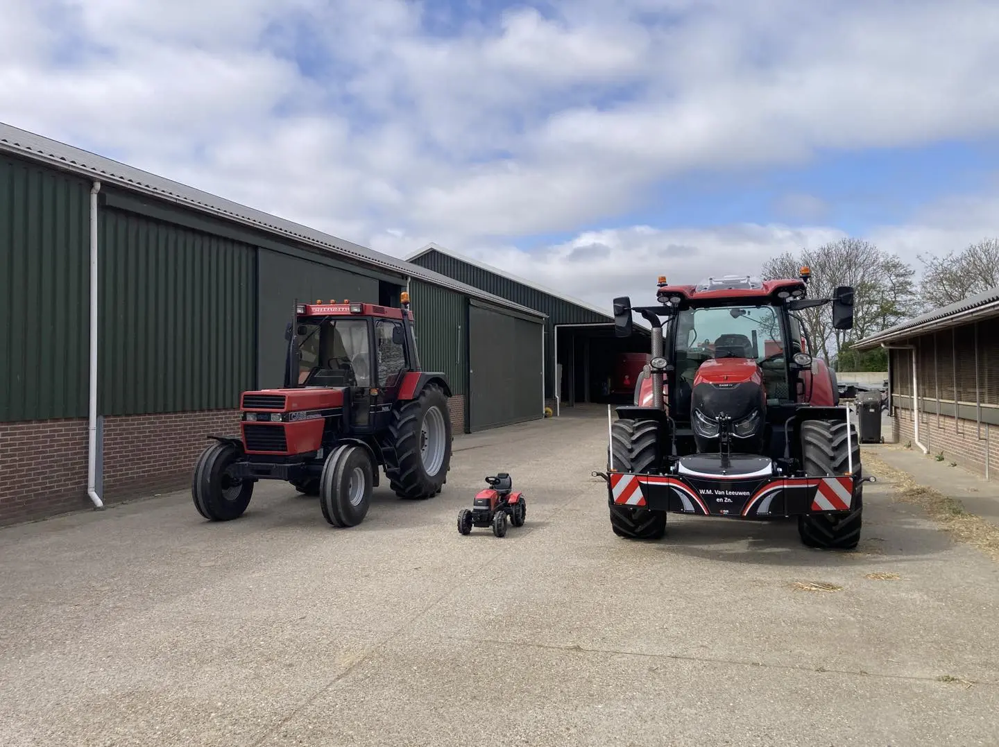 Two red tractors and a small red ride-on toy tractor are parked on a concrete yard beside green farm buildings