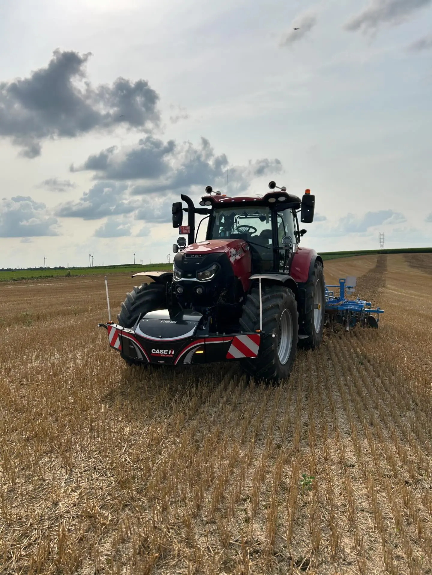 Red tractor with a front weight bumper and striped hazard markings in a harvested field under a cloudy sky
