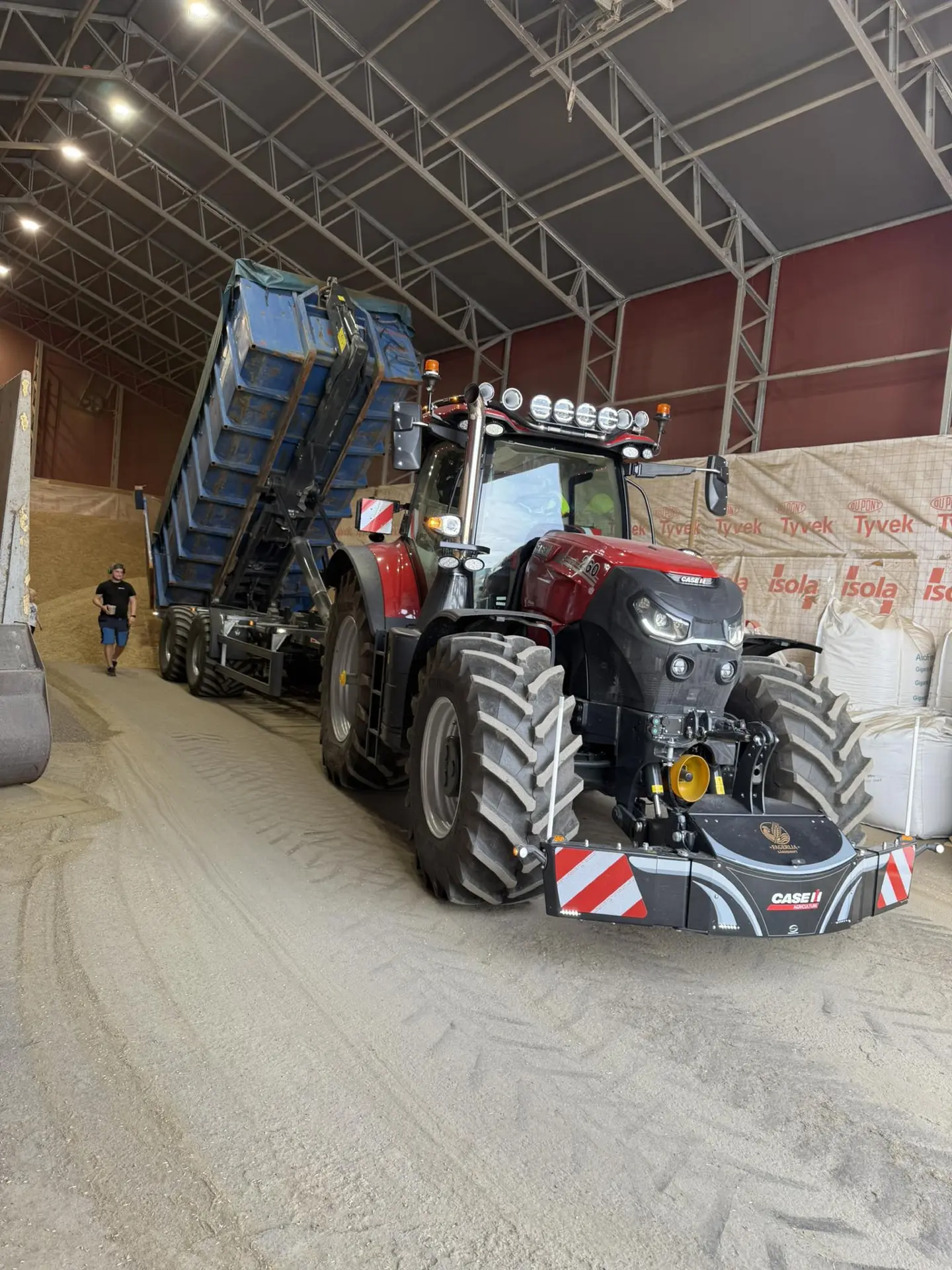 Red tractor with a front weight bumper and a raised blue trailer inside a large warehouse with a person nearby