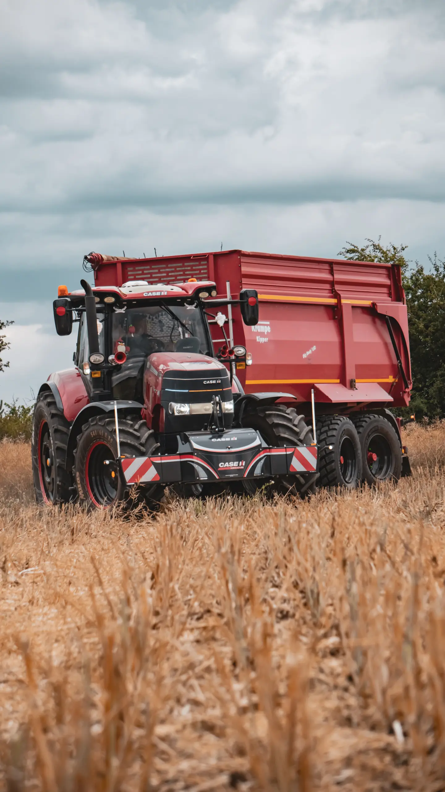 Red tractor with a front weight bumper and a red trailer driving through a harvested field under a cloudy sky