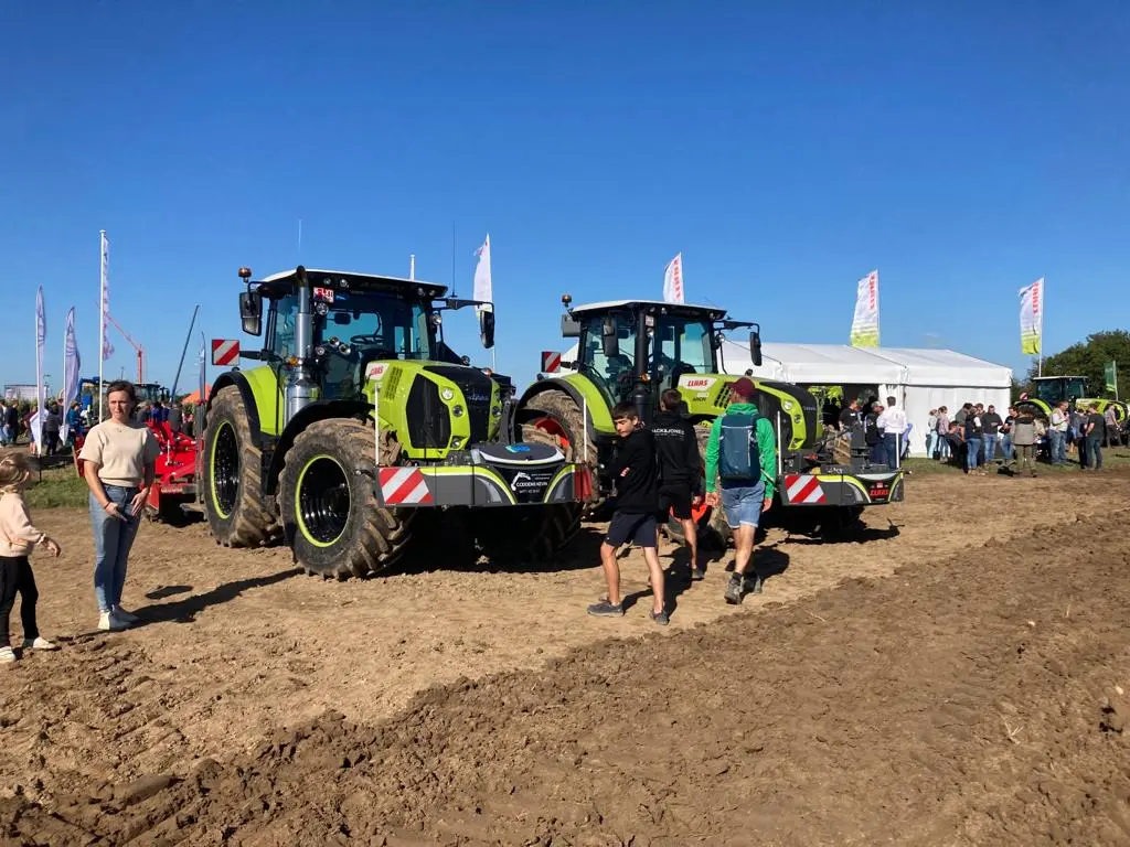Two green tractors with front weight bumpers on display outdoors with people walking around on a sunny day