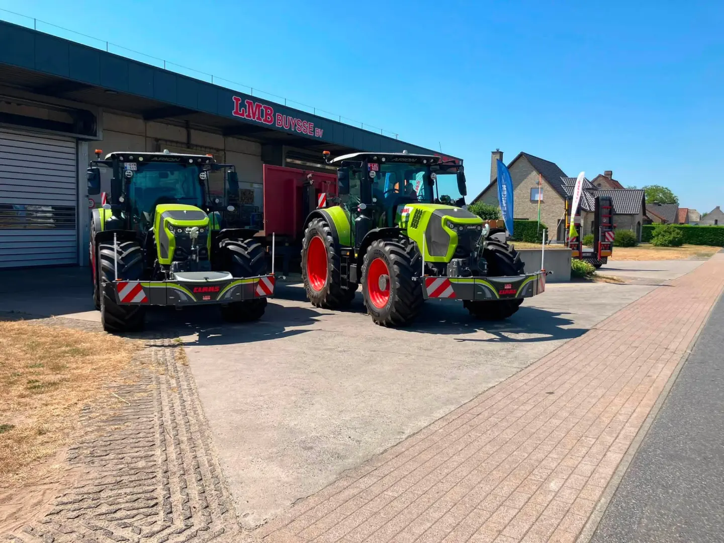 Two green tractors with front weight bumpers are parked on concrete outside a building under a blue sky