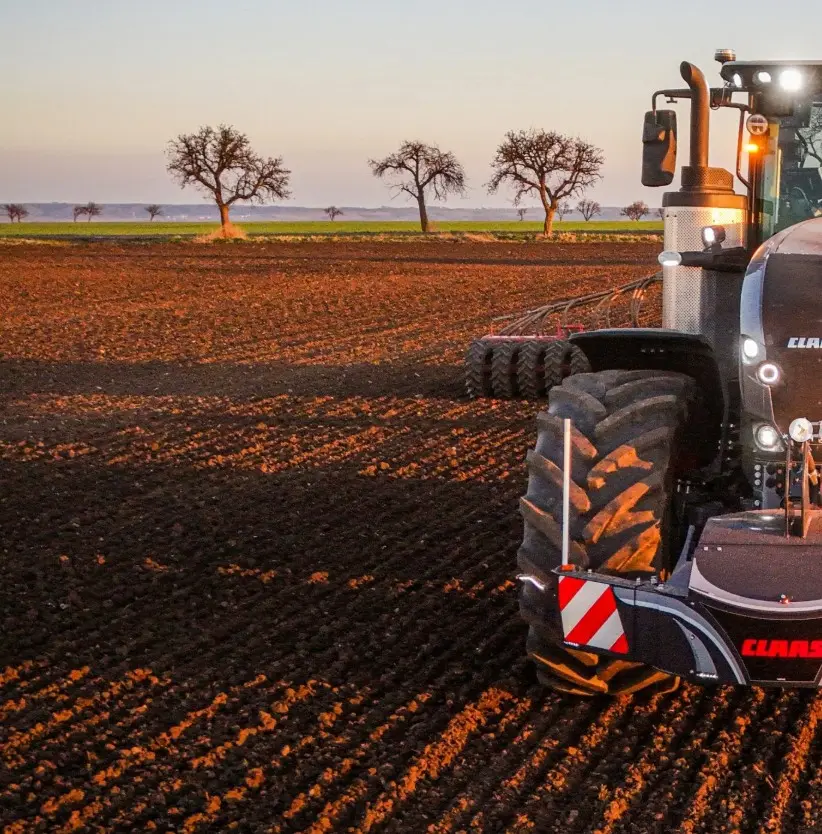 Front view of a tractor with a CLAAS-branded front weight bumper and LED lights in a plowed field at sunset