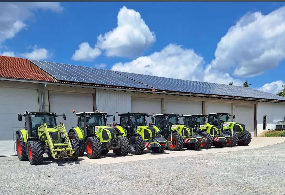 Six green tractors with front weight bumpers are parked in a row outside a building with solar panels
