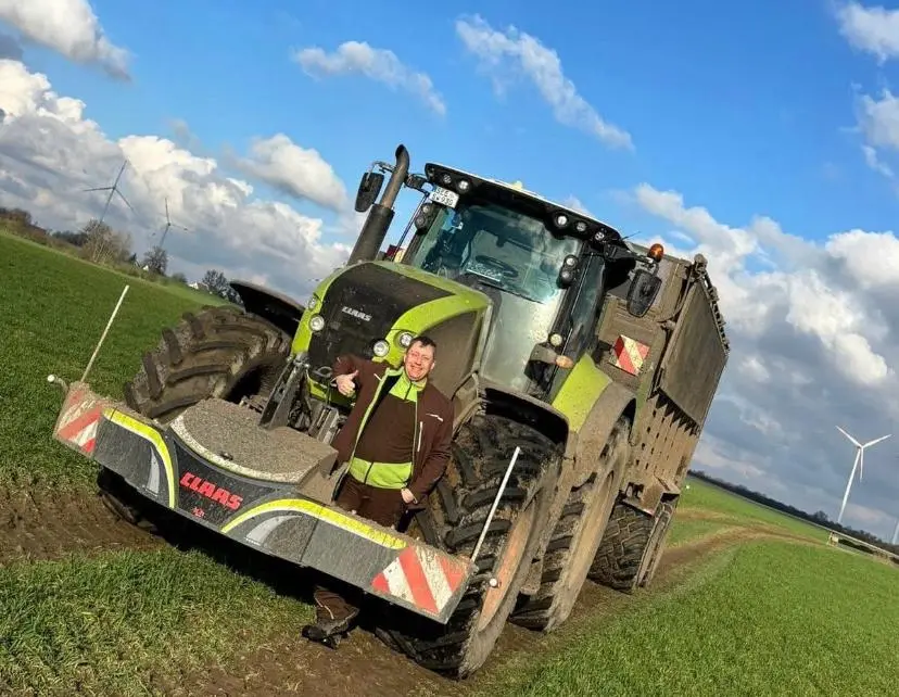 A person stands in front of a tractor with a CLAAS front weight bumper on a grassy field under a blue sky.