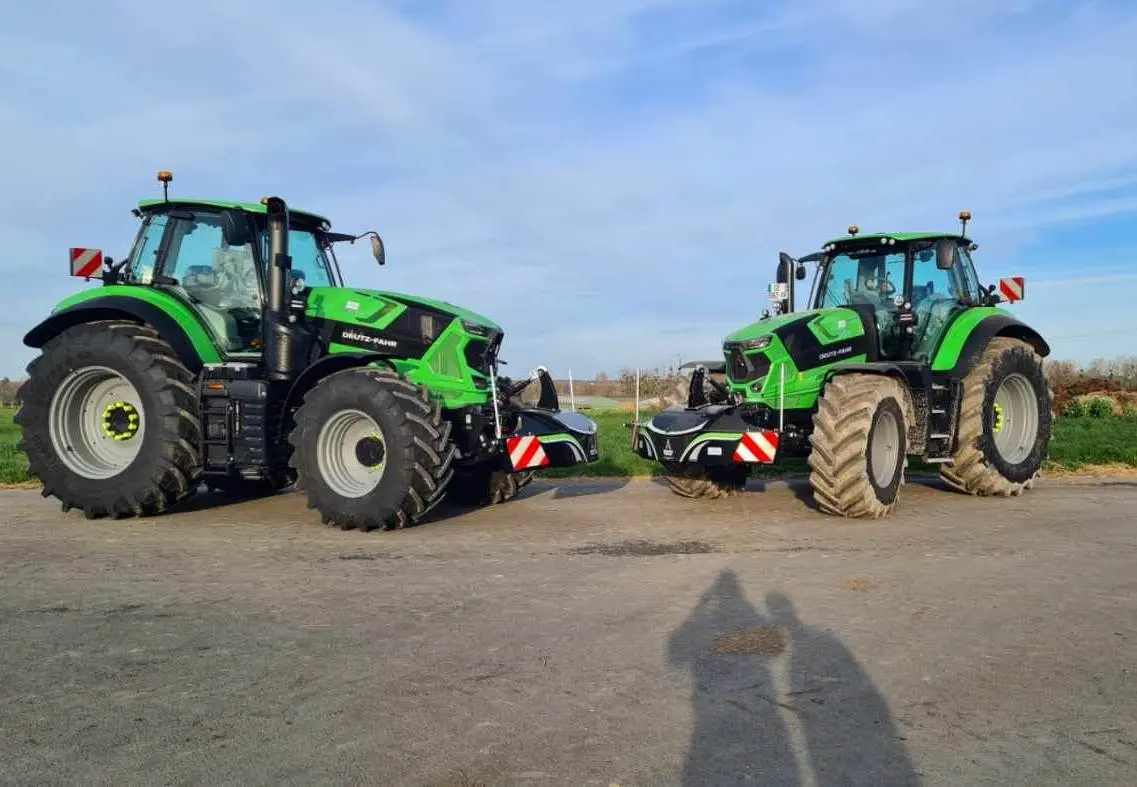 Two green tractors with mounted front weight bumpers and red-white safety markings are parked on a farm road