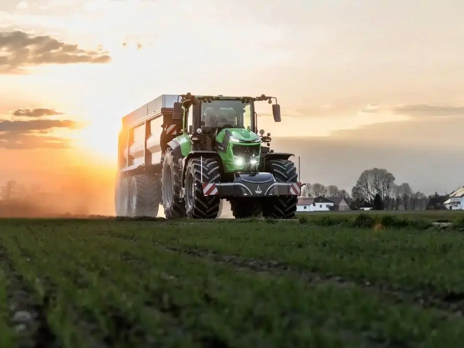 A green tractor with a front weight bumper is driving on a field at sunset with a trailer attached