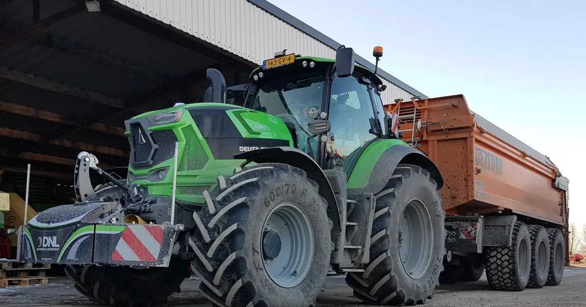 Green tractor with a front weight bumper and large trailer parked near a shed on a farmyard