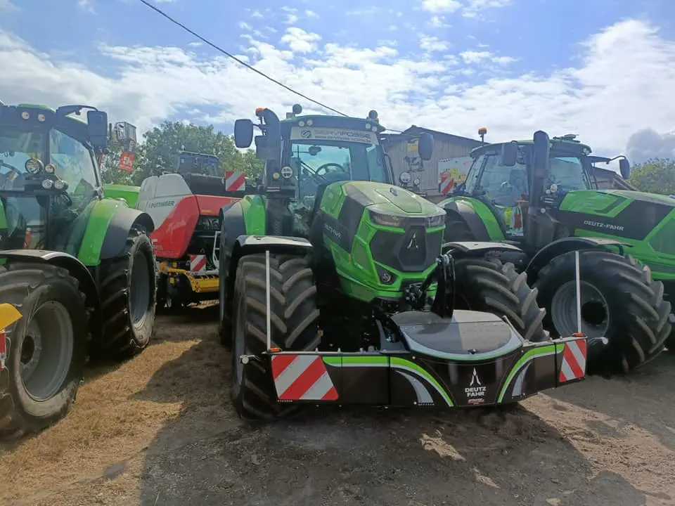 Green tractor with a large black and silver front weight bumper, parked among other tractors on a sunny day