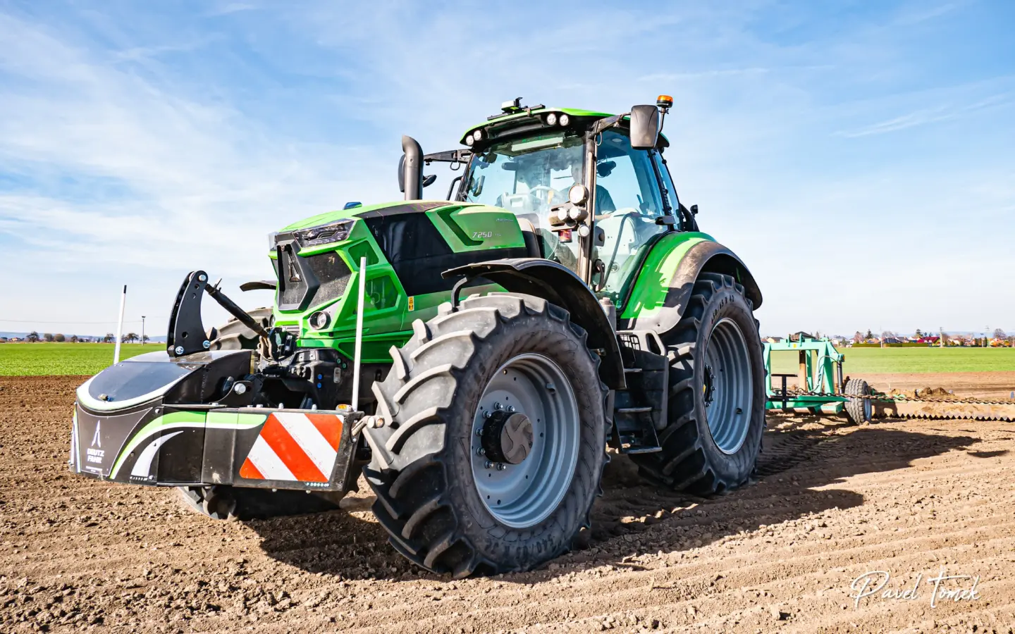 Green tractor with a large front weight bumper attached, parked on tilled soil in a field under a blue sky