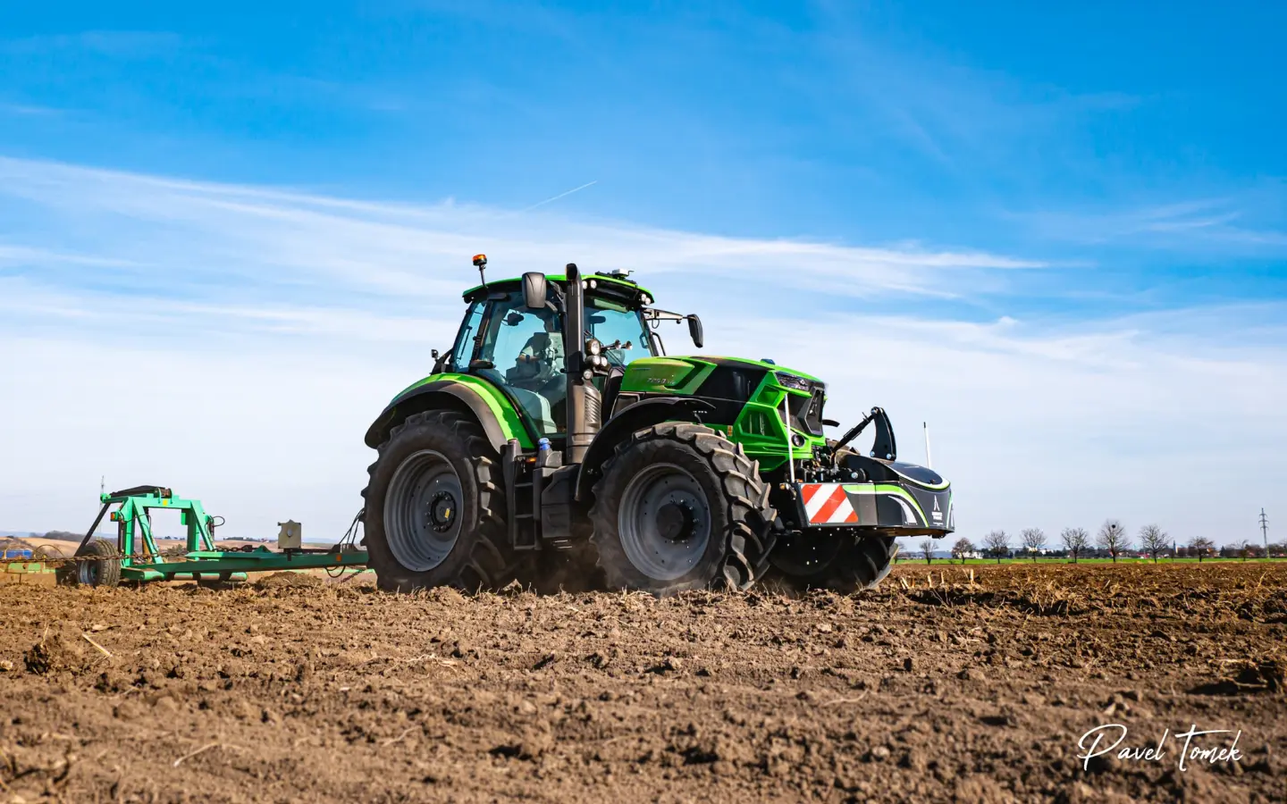 Green tractor with a large front weight bumper working in a plowed field under a blue sky