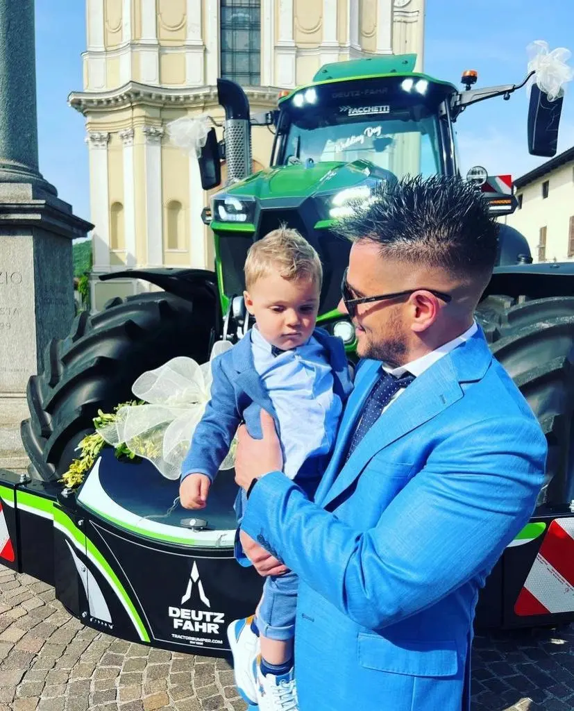 A man holding a child stands in front of a Deutz-Fahr tractor with a front weight bumper decorated with a large bow