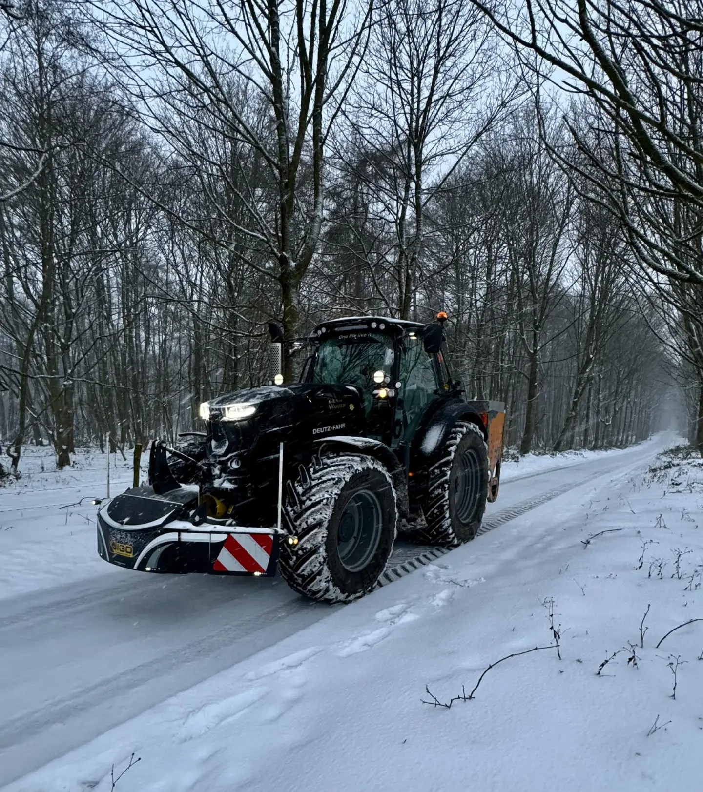 A tractor fitted with a front weight bumper drives on a snowy road through a forested area