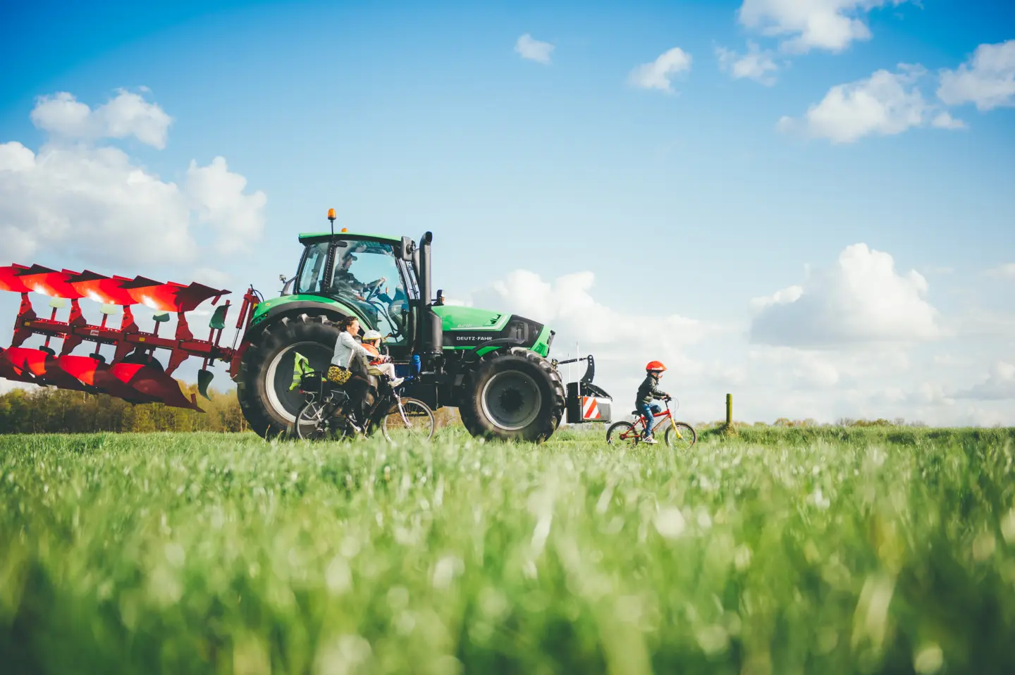 A green tractor with a front weight bumper and red tillage equipment in a grassy field, near a woman and children on bicycles