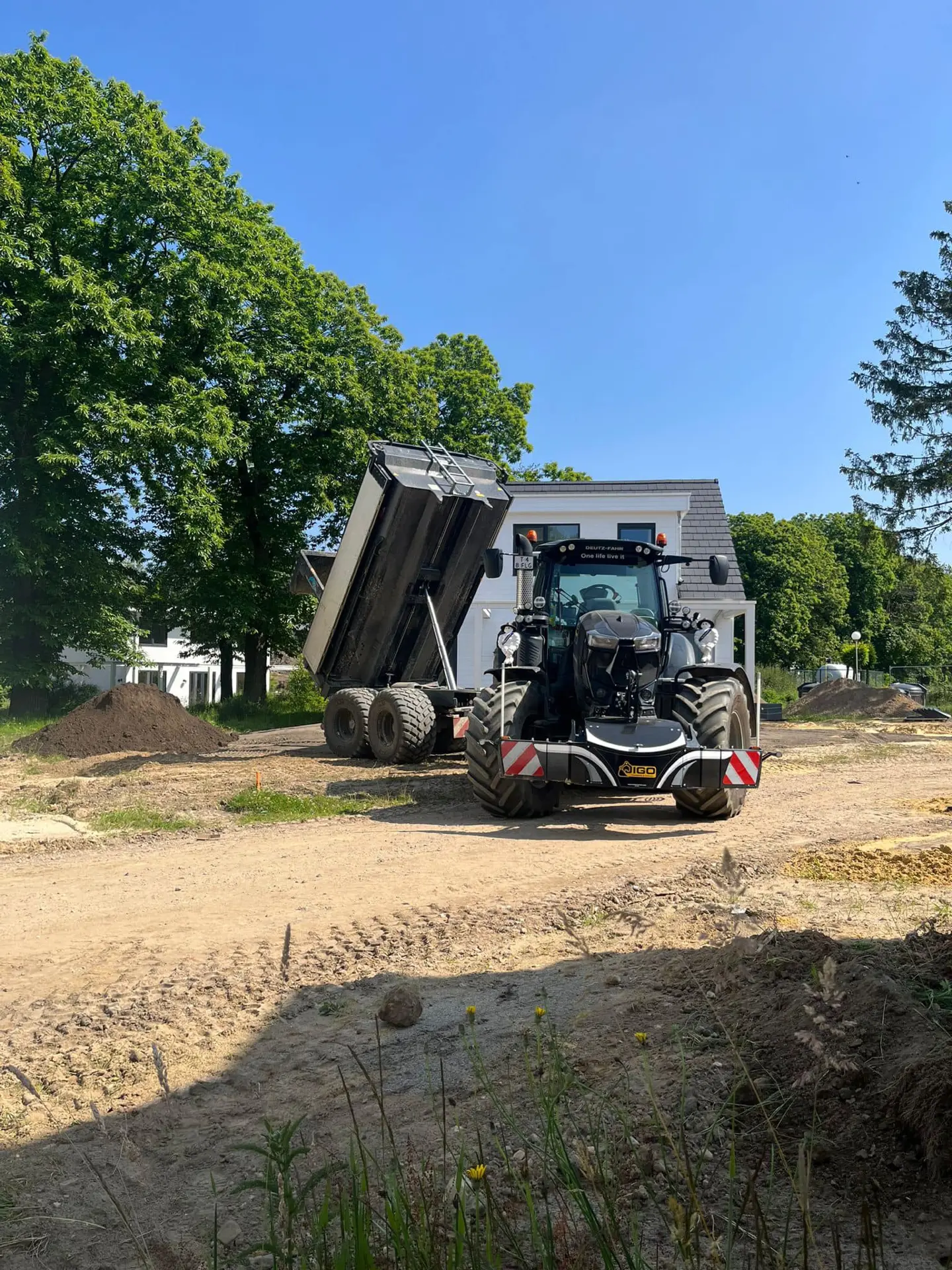 A tractor with a front weight bumper is parked on a construction site beside a trailer with its bed raised