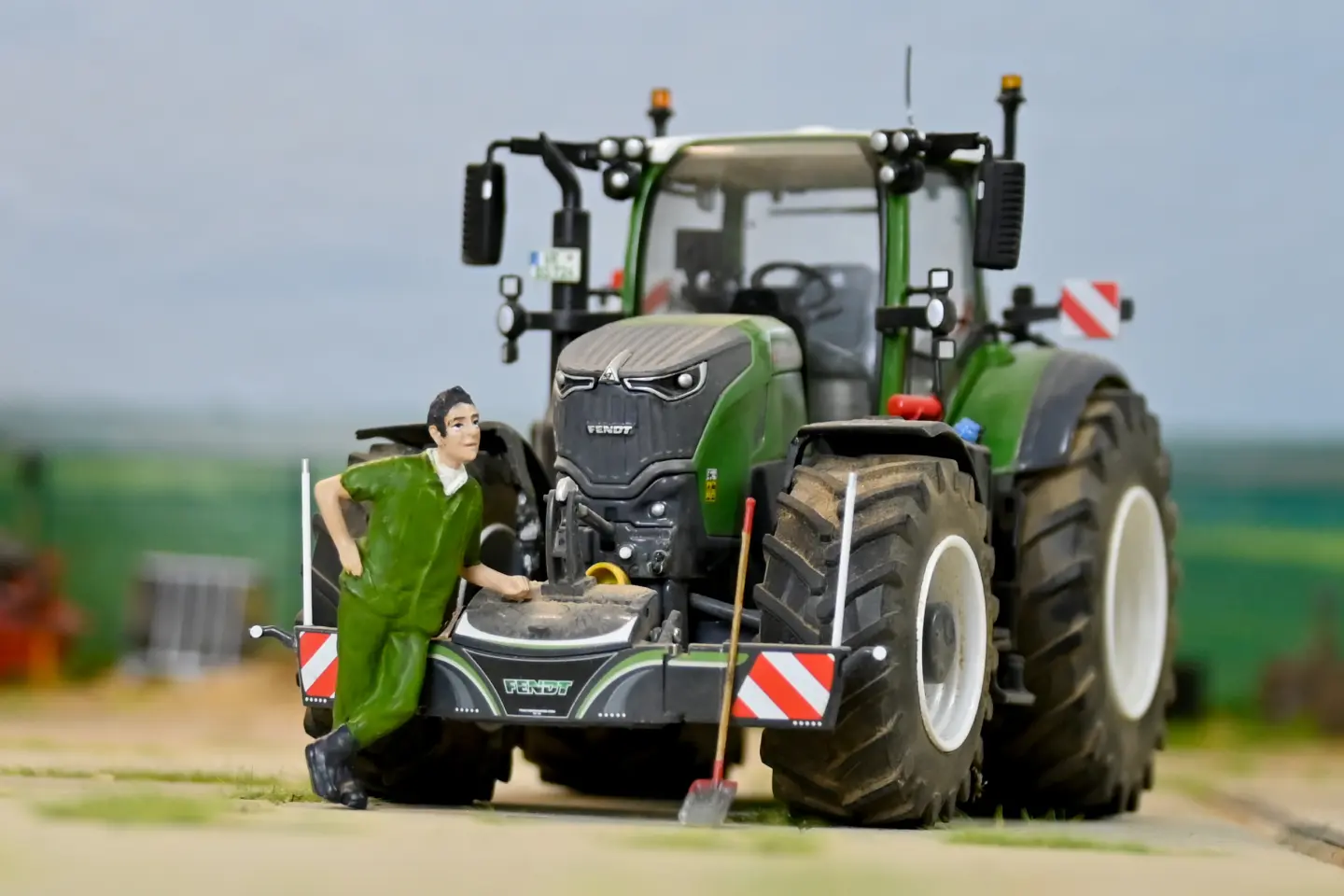 A model tractor with a front weight bumper, warning stripes and a figurine in green overalls leaning on it