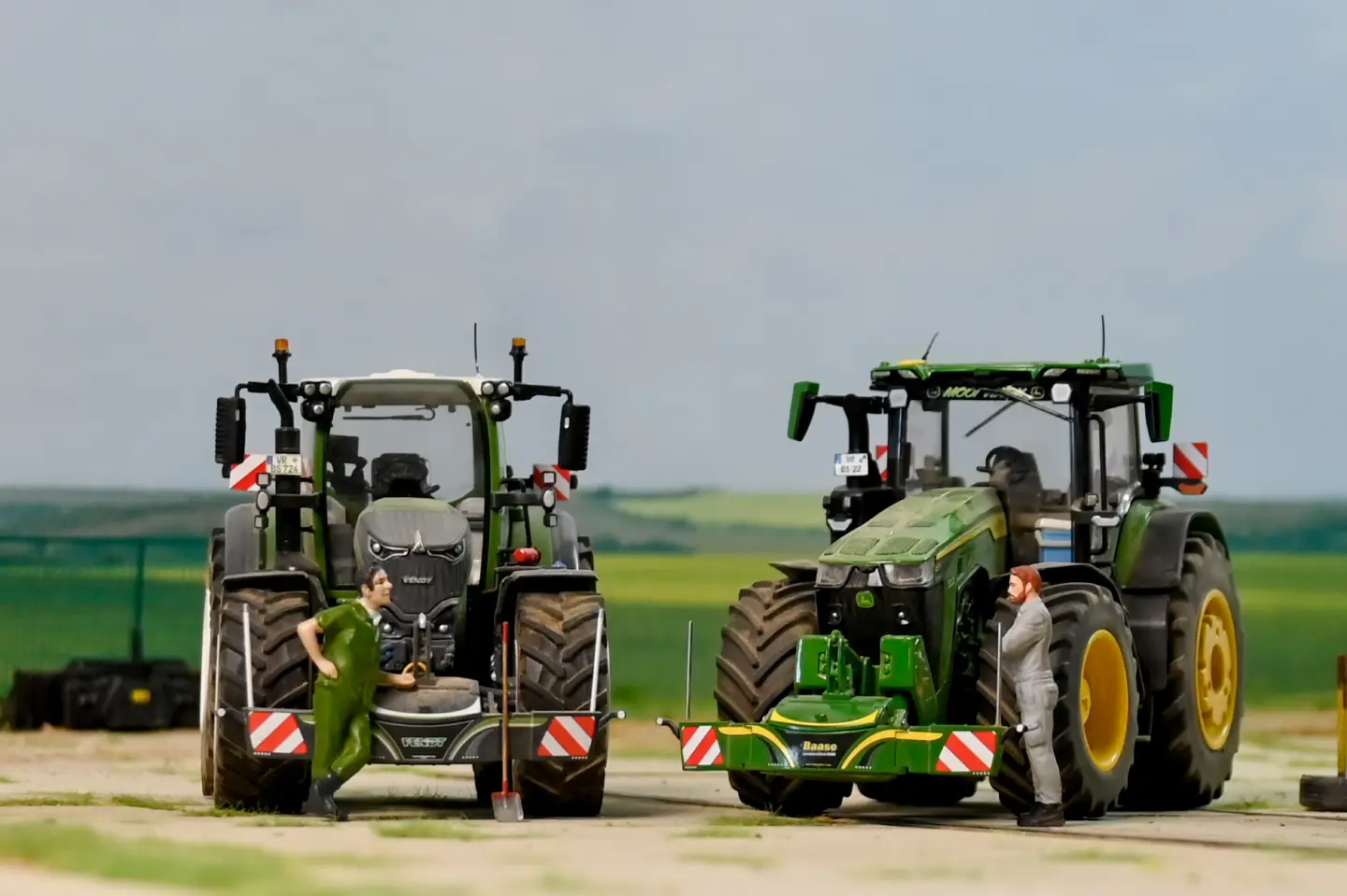 Two tractors with front weight bumpers are parked on a dirt surface, each with a person standing nearby.