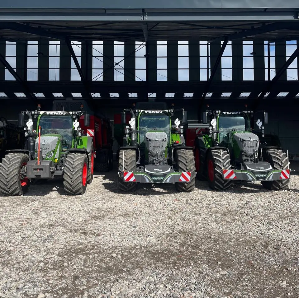 Three green tractors with front weight bumpers and red wheel rims are parked side by side on gravel under a shed