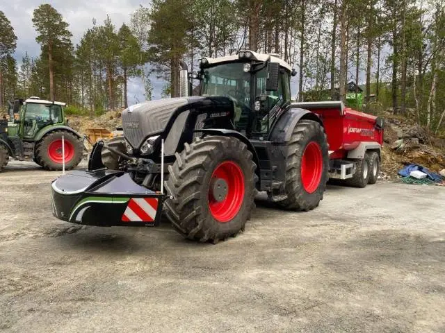 Black tractor with a front weight bumper attached, pulling a red trailer, parked outdoors on a dirt surface