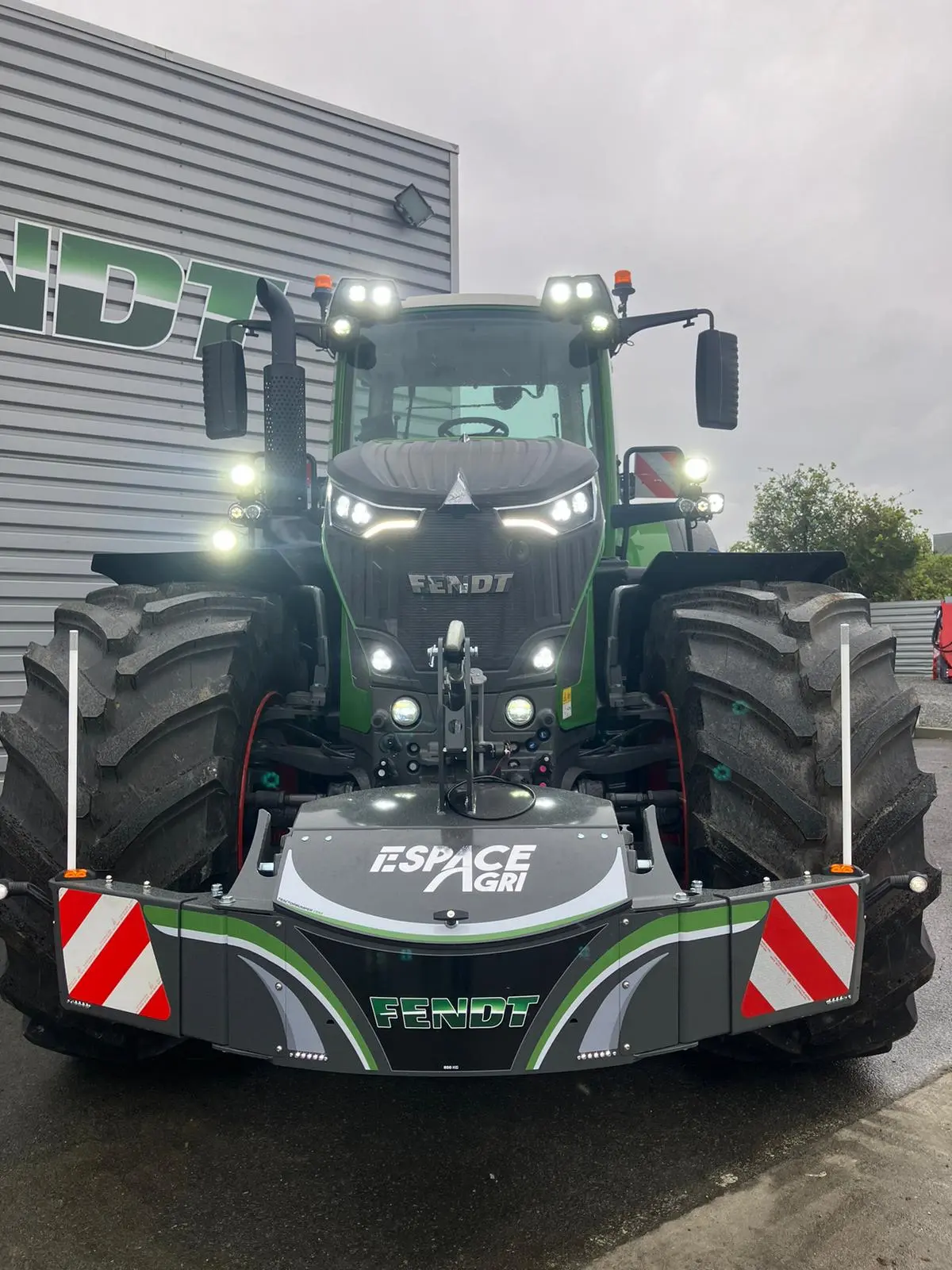 Large tractor with a front weight bumper featuring Espace Agri and Fendt branding, parked outside a building