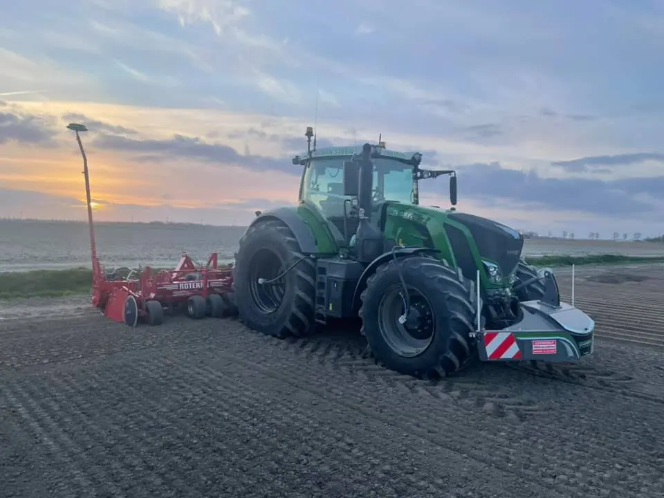 A green tractor with a front weight bumper and red agricultural attachment works in a plowed field at sunset