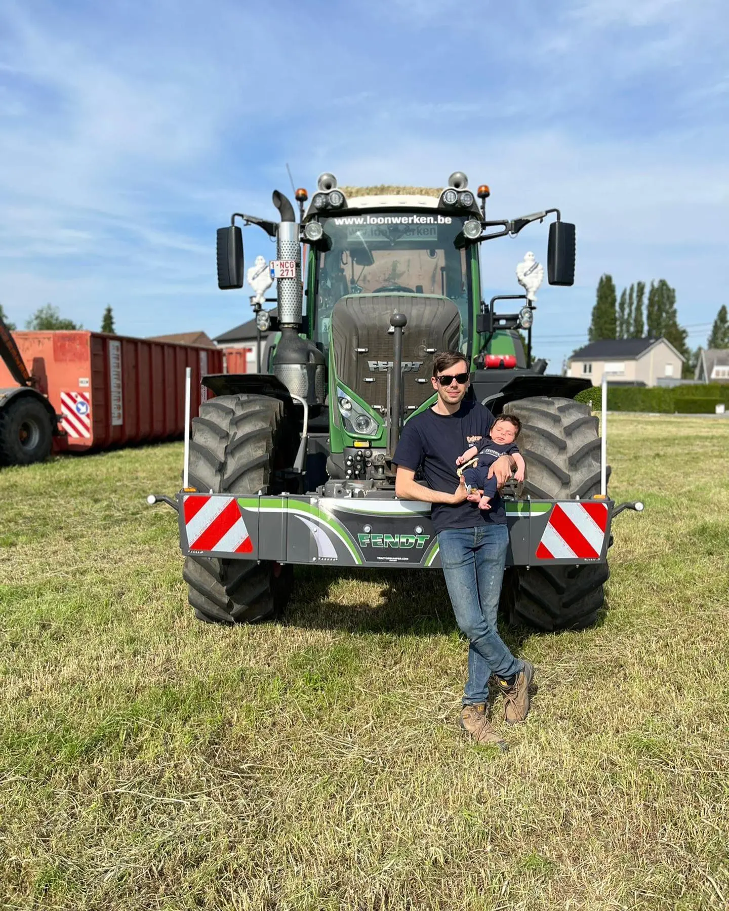 A man holding a baby stands in front of a tractor fitted with a front weight bumper on a grassy field