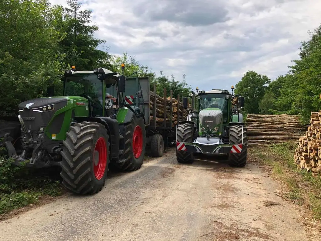 Two green tractors with front weight bumpers are parked on a rural road, each hauling trailers loaded with logs