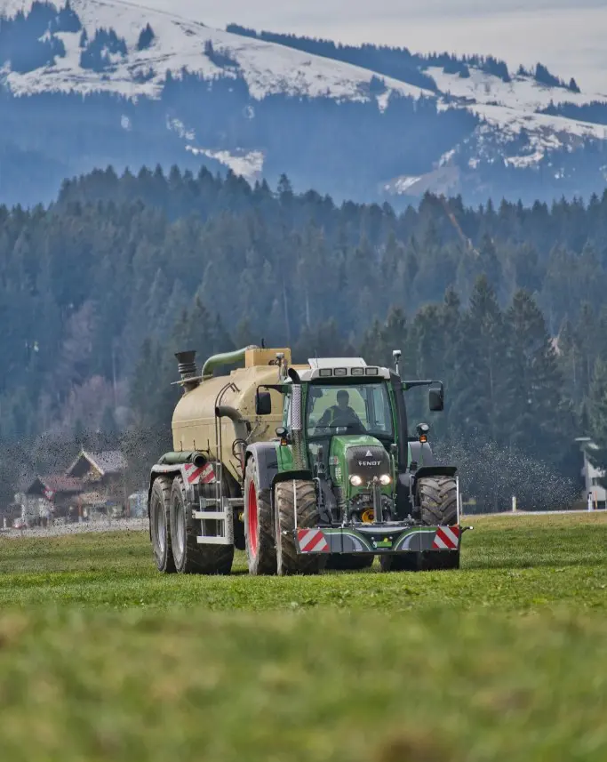 A green tractor with a front weight bumper and attached tan tanker drives across a grassy field with mountains in the