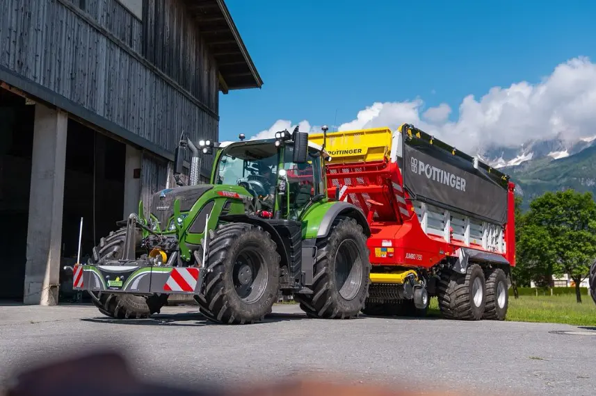 A green tractor with a front weight bumper is attached to a large red trailer parked outdoors near a barn