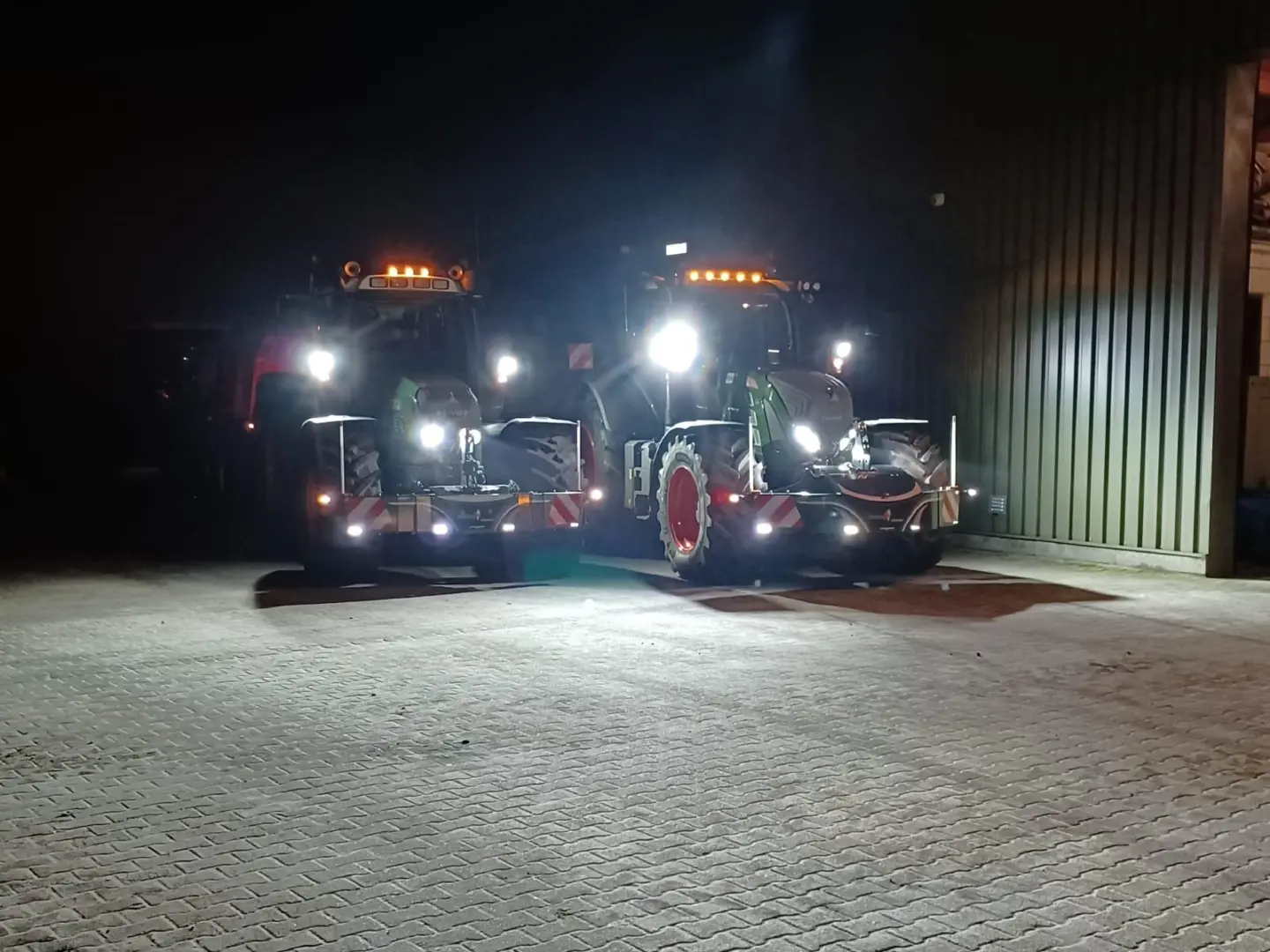 Two tractors with front weight bumpers and headlights on are parked outside a building at night
