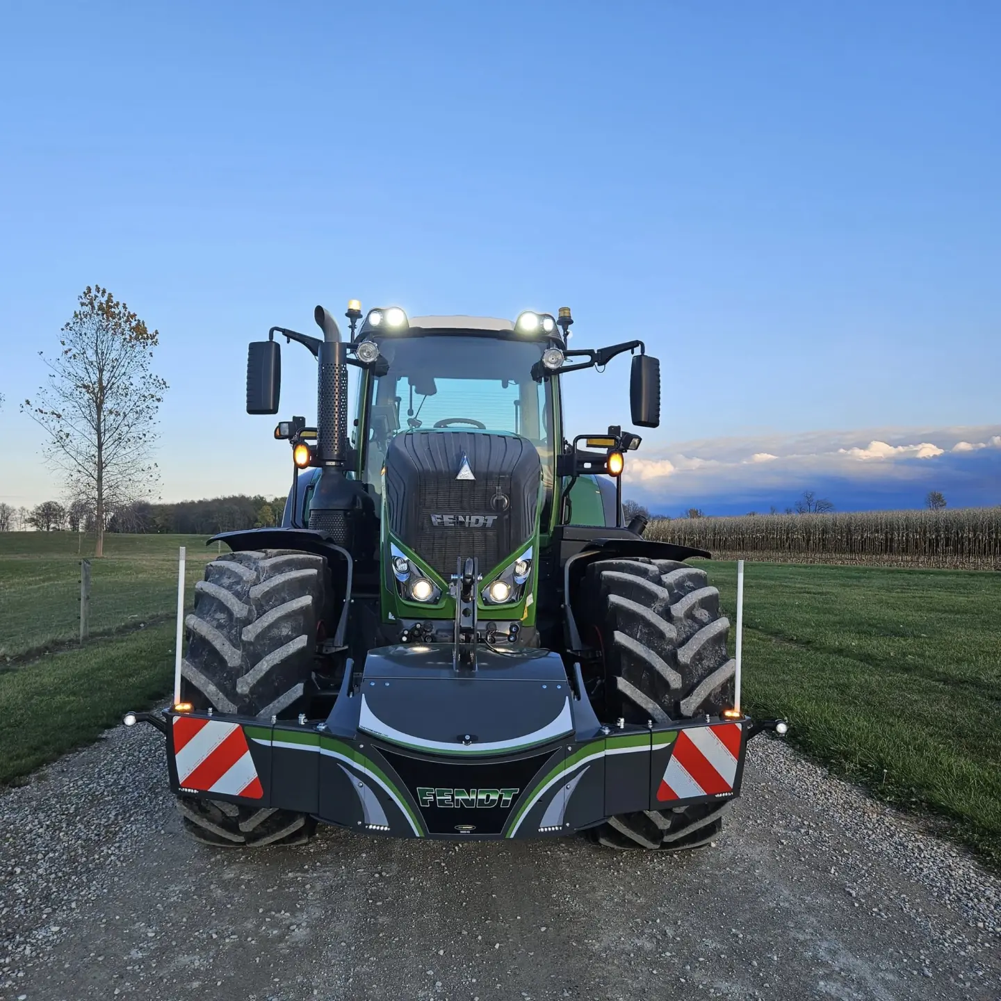 Green tractor with large tires and a front weight bumper, parked on a gravel road with warning stripes visible