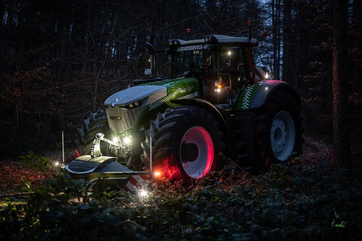 Green tractor with illuminated front weight bumper attachment on a forest path at dusk, lights glowing warmly