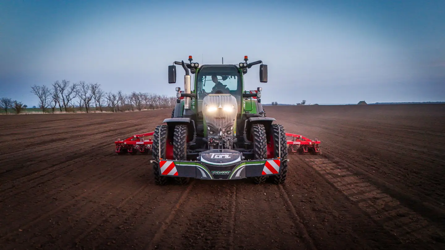 A tractor with headlights on, fitted with a front weight bumper, works a field at dusk with red equipment attached