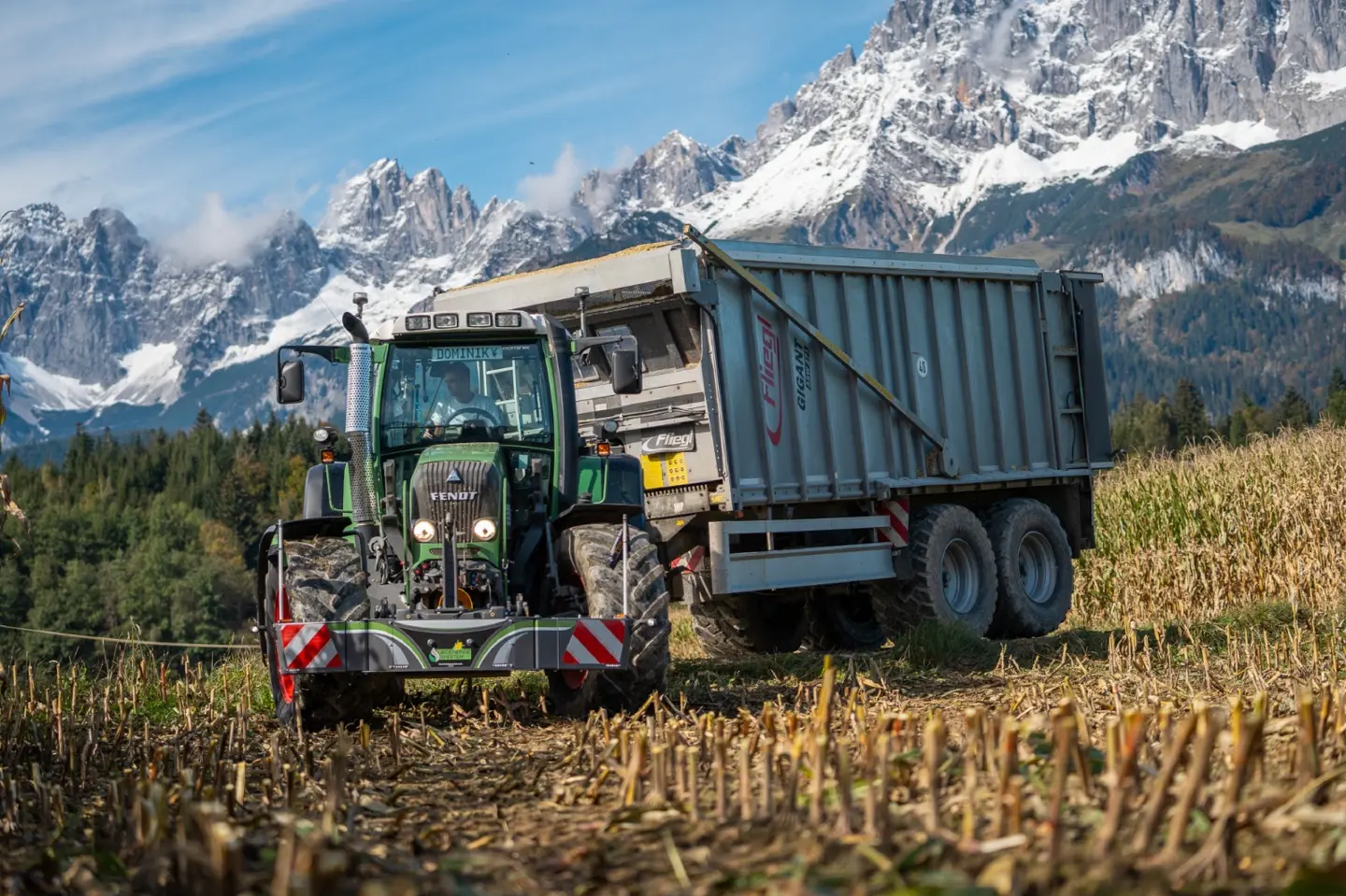 A tractor with a front weight bumper and attached trailer is working in a harvested field with mountains in the background