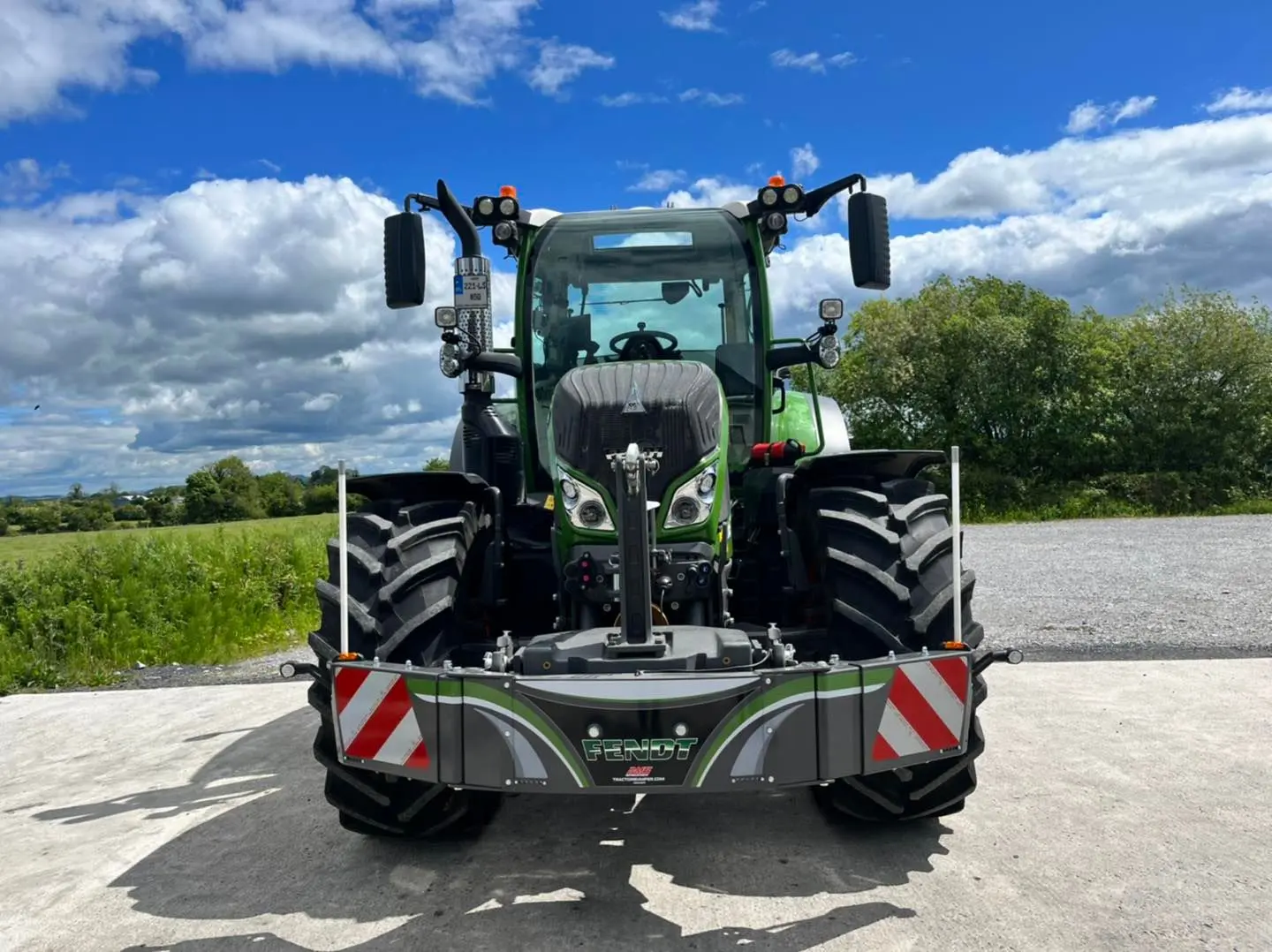 A green tractor with a Fendt-branded front weight bumper is parked on concrete near a grassy field under a blue sky
