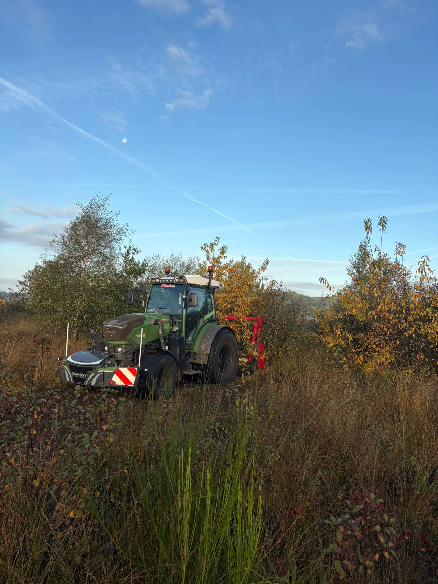 Green tractor with front weight bumper and rear attachment parked in tall grass with trees and blue sky in the background