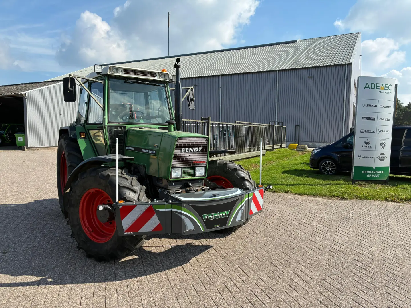 A green tractor with a front weight bumper is parked on a paved area near a building and a sign with company logos