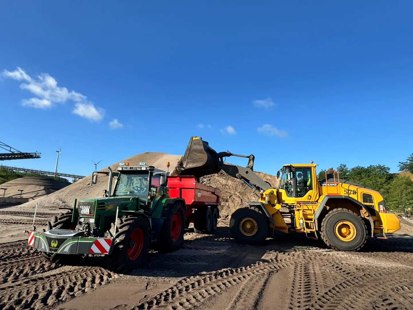 A green tractor with a front weight bumper and red trailer is parked beside a yellow wheel loader at a sandy worksite