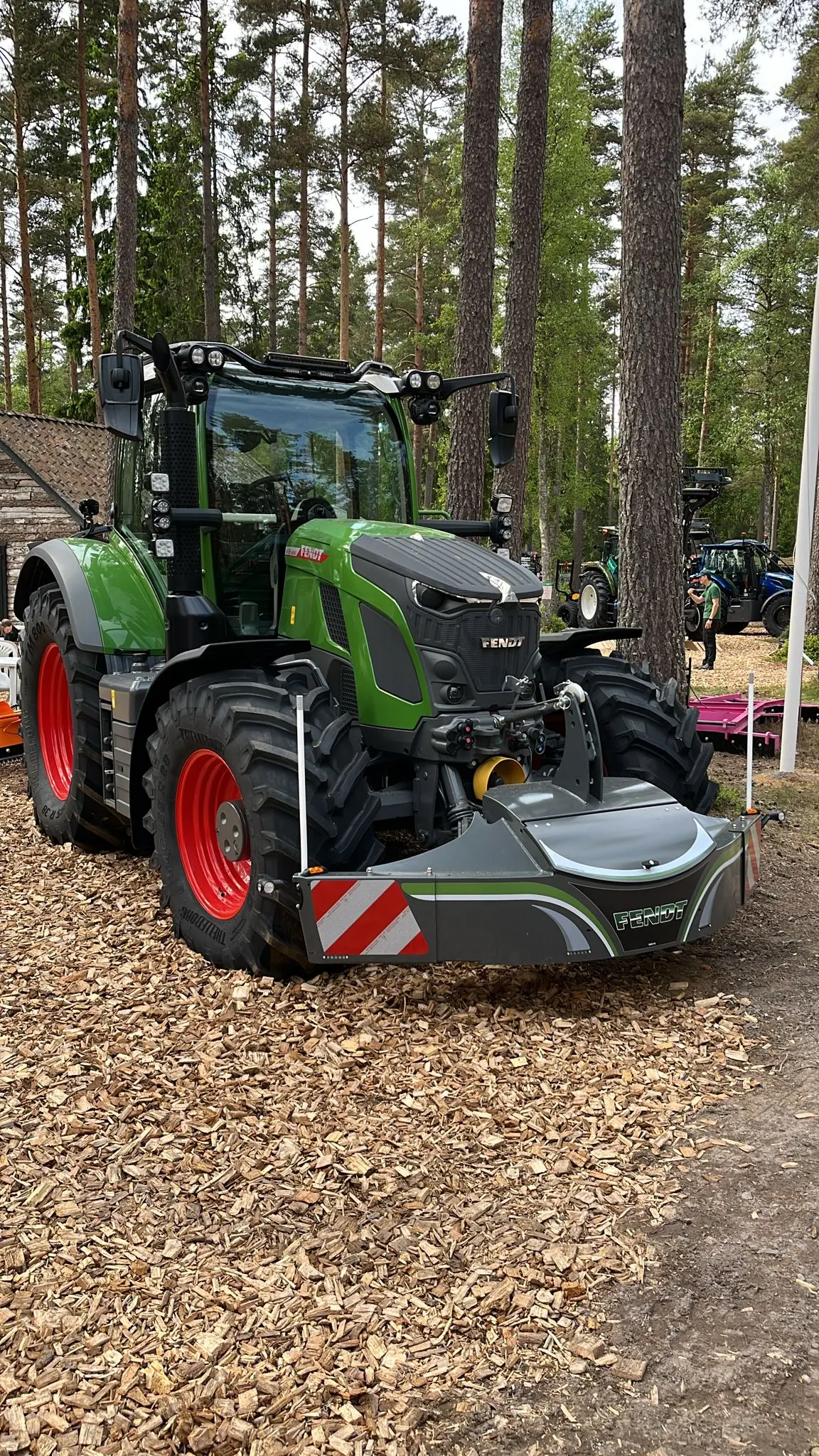 A green tractor with large red wheels fitted with a Fendt-branded front weight bumper in a wooded outdoor area