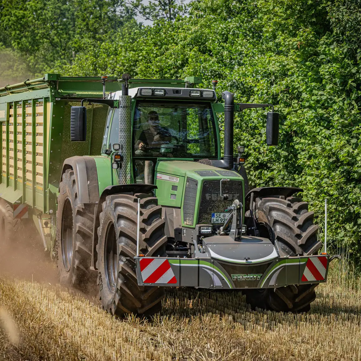 Green tractor with a front weight bumper attached, pulling a large trailer through a field near tall green hedges