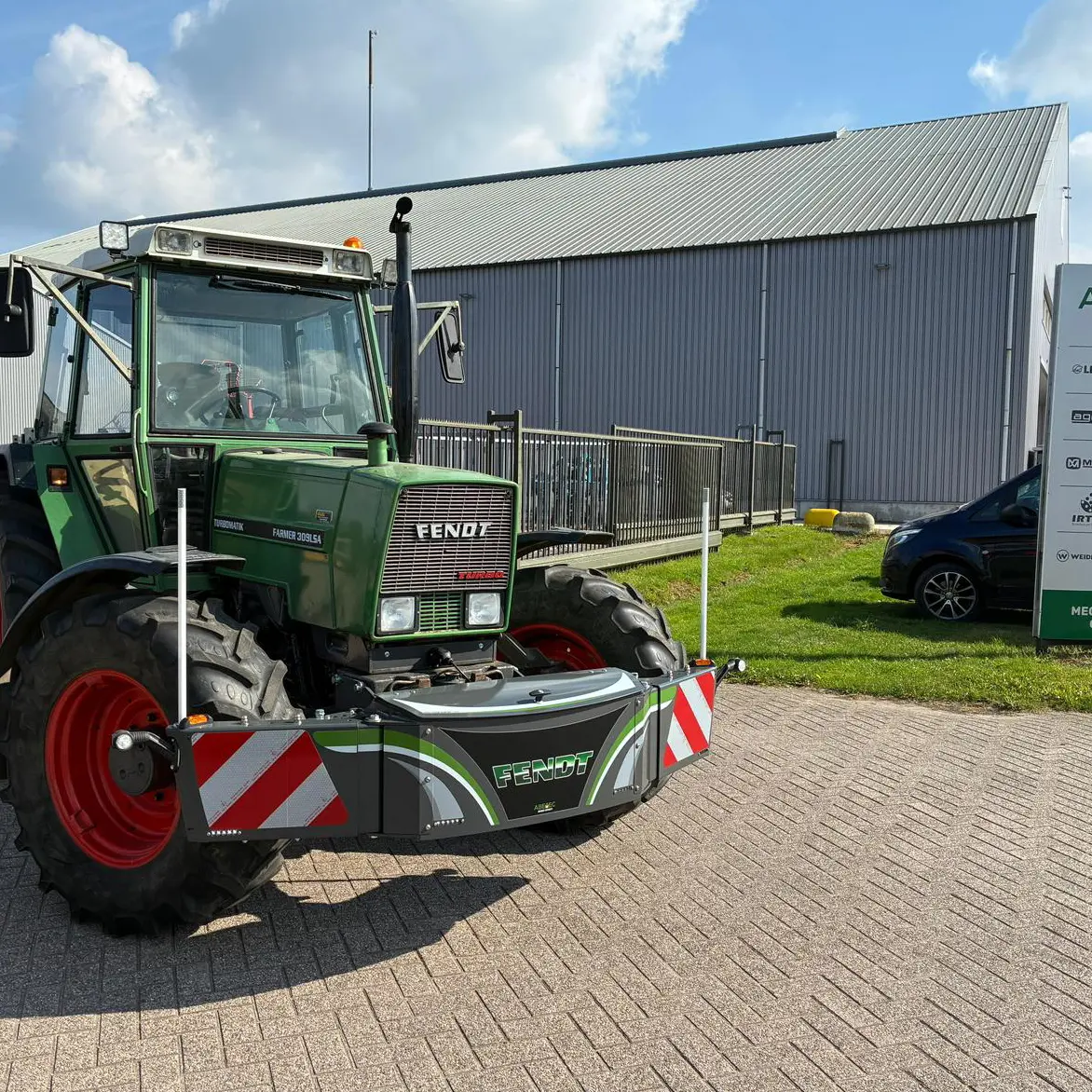 Green tractor with a front weight bumper attached, parked on a paved area near a metal building and sign