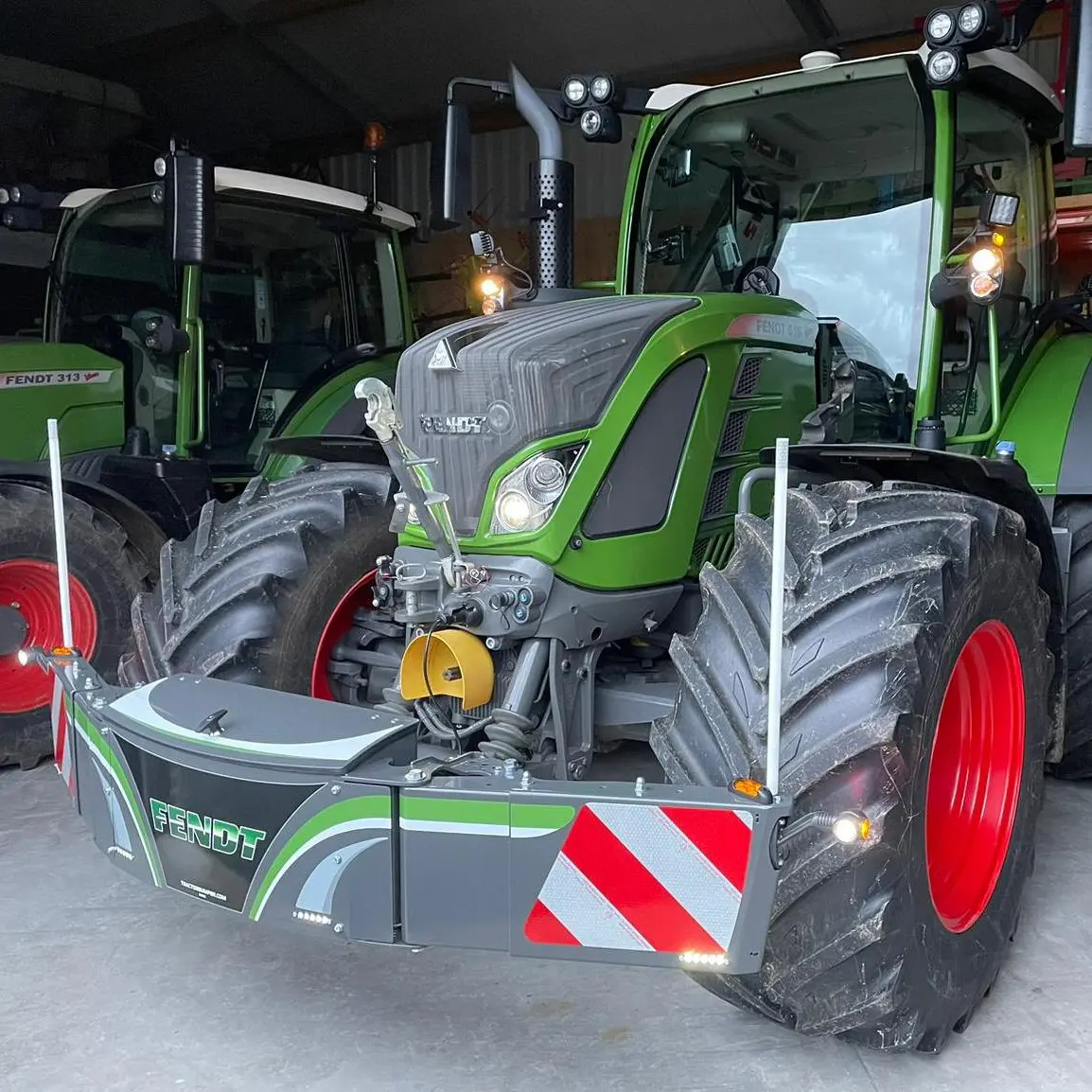 Green tractor with a front weight bumper featuring FENDT branding, reflective panels and guide poles in a shed