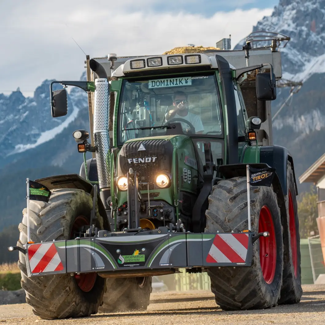 Green tractor with large front weight bumper featuring reflective red and white stripes, driving outdoors on gravel