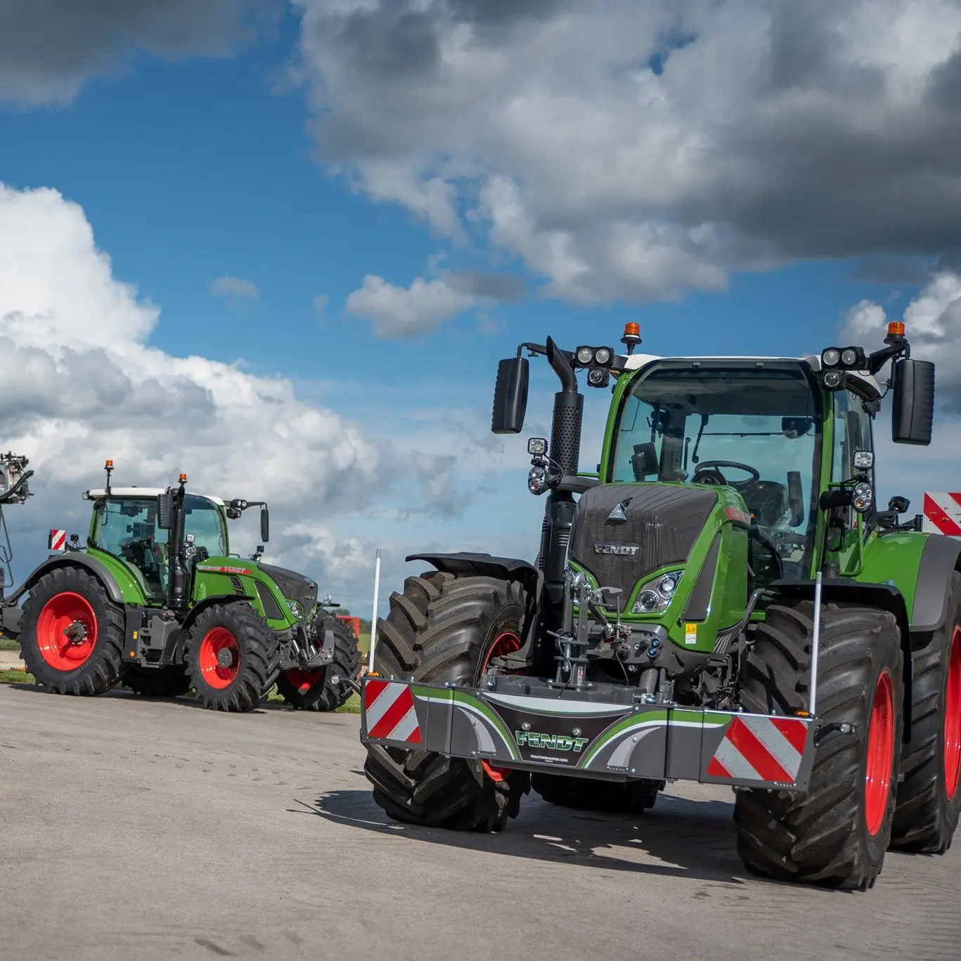 Two green tractors with front weight bumpers and orange wheels are parked outdoors under a partly cloudy sky